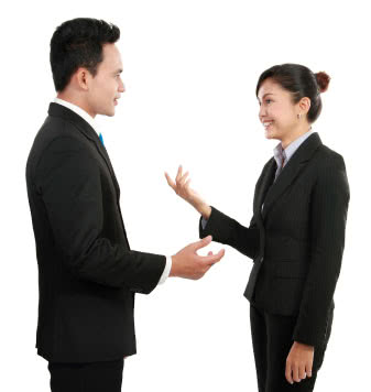 A male in a suit with his hand in the position of a shake, questioning a female in a suit who appears to be answering, her left arm slightly lifted