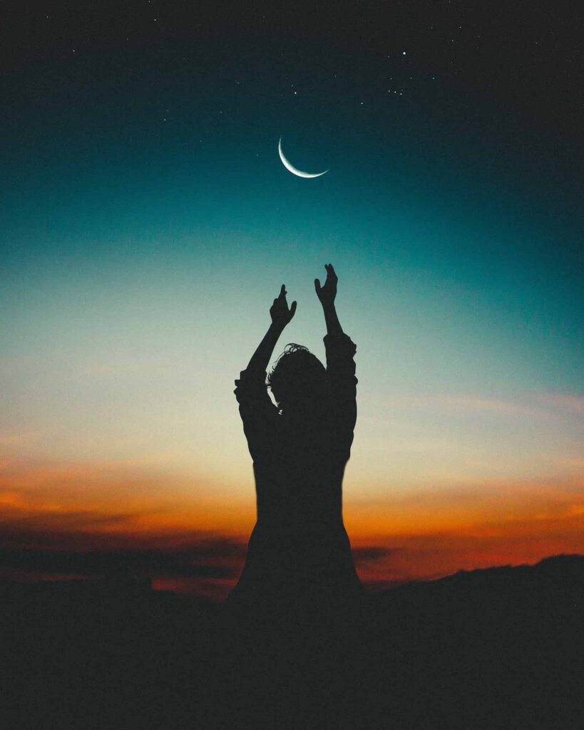 Silhouette of person reaching for crescent moon, and the time of day is dusk with vibrant orange, blue, and black in background
